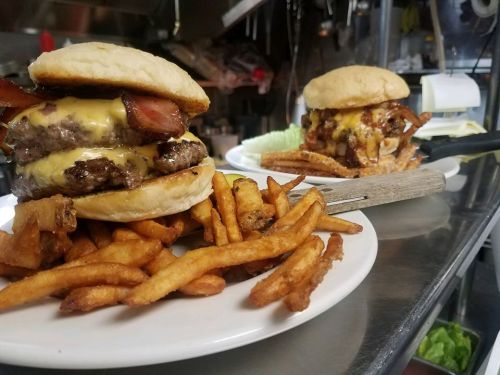 Two hearty burgers with melted cheese and bacon, served with a side of crispy fries on a stainless steel countertop.