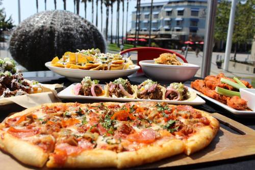 A variety of delicious dishes including pizza, nachos, tacos, and wings, with palm trees and a building in the background.