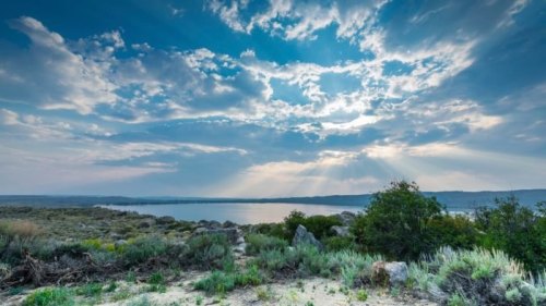A scenic view of a lake surrounded by greenery, under a dramatic sky with rays of sunlight breaking through the clouds.