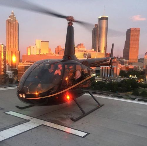 A helicopter on a rooftop helipad with a city skyline at sunset in the background.