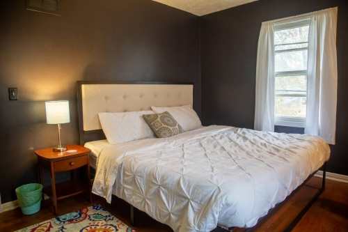 Cozy bedroom with a queen bed, nightstand, lamp, and a window with sheer curtains against dark walls.