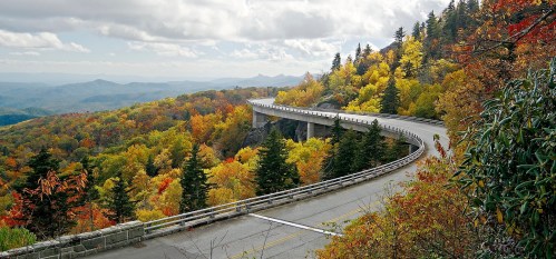 A winding road curves through vibrant autumn foliage on a mountain landscape under a cloudy sky.