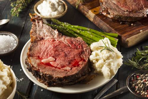 A plate of juicy prime rib with mashed potatoes, asparagus, and sauces, set on a rustic wooden table.