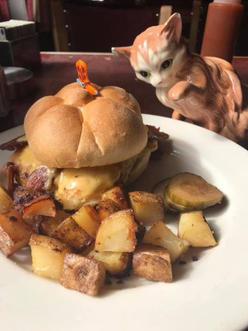 A close-up of a cheeseburger with bacon on a bun, served with roasted potatoes, and a ceramic cat figurine in the background.