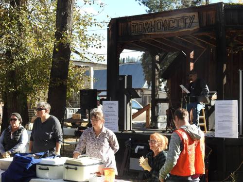 A group of people at a community event in Idaho City, with a stage and trees in the background.