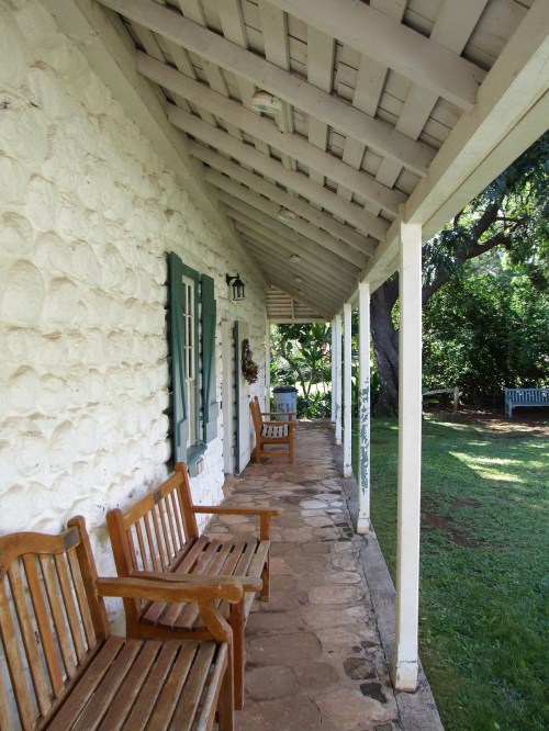 A porch with wooden benches, stone walls, and a sloped roof, surrounded by greenery and a peaceful garden.