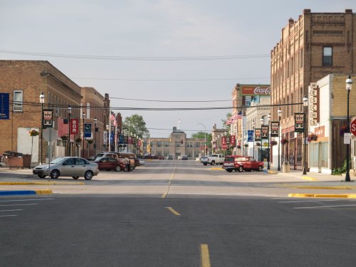 A quiet downtown street lined with brick buildings, flags, and parked cars under a clear sky.