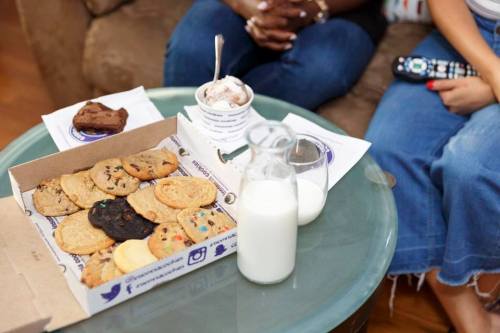 A box of assorted cookies, a glass of milk, and ice cream on a table, with two people sitting nearby.
