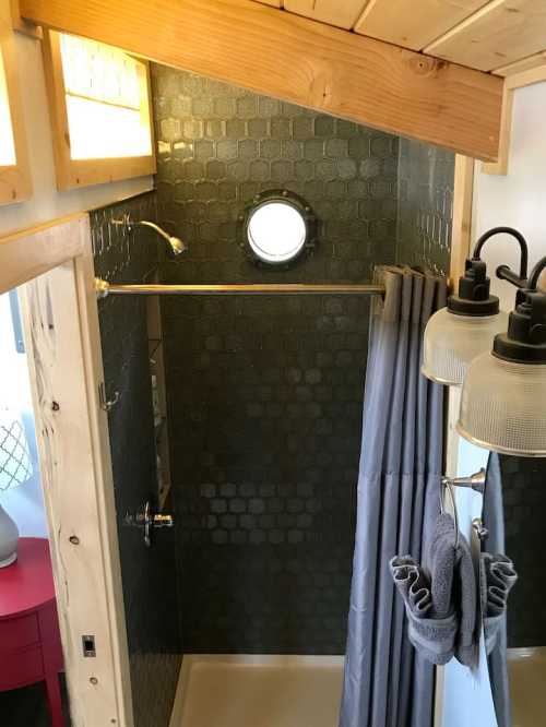 A compact shower area with green hexagonal tiles, a circular window, and gray curtains in a wooden interior.