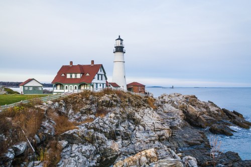 A lighthouse stands on rocky shores, beside a charming house, with calm waters and a cloudy sky in the background.
