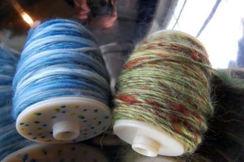 Two spools of yarn: one blue with white stripes and one multicolored in green, red, and brown, resting on a reflective surface.