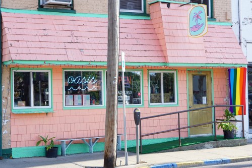 A pink building with a sign reading "Oasis," featuring a rainbow flag and green plants outside.