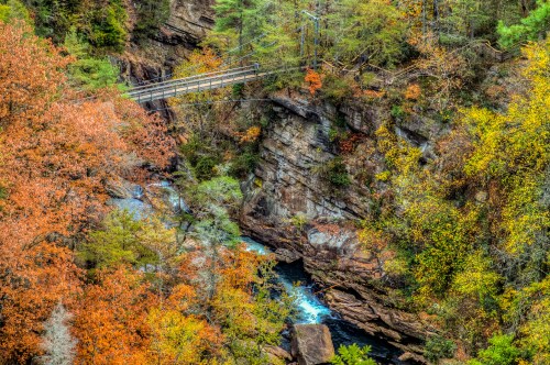 A scenic view of a suspension bridge over a river, surrounded by vibrant autumn foliage and rocky cliffs.