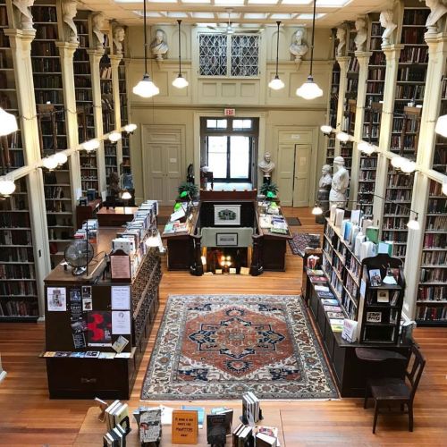 A spacious library with tall bookshelves, wooden floors, and decorative statues, featuring a central reading area.
