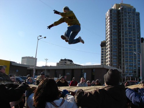 A person in a yellow jacket leaps through the air above a crowd holding a large tarp, with buildings in the background.