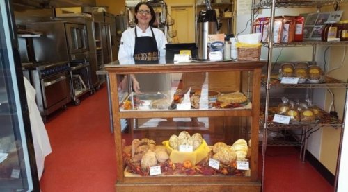 A smiling baker stands behind a glass display case filled with various baked goods in a cozy bakery.
