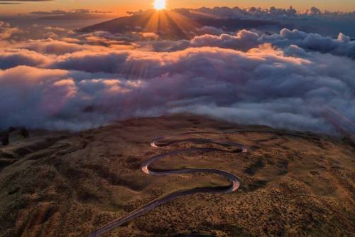 A winding road curves through a landscape above the clouds, with a vibrant sunset illuminating the scene.