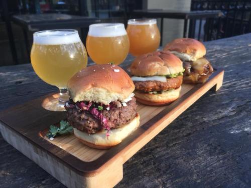 A wooden platter with three mini burgers and four glasses of light beer on a rustic table.