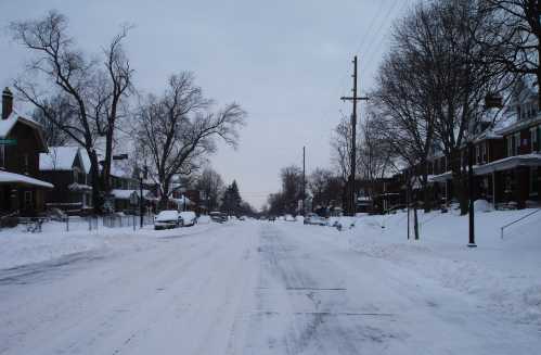 A snowy street lined with houses, trees, and parked cars, under a gray sky.