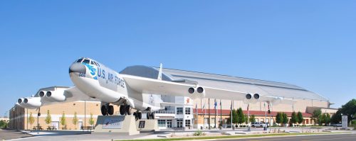 A large aircraft displayed outside a modern building, with flags and clear blue sky in the background.