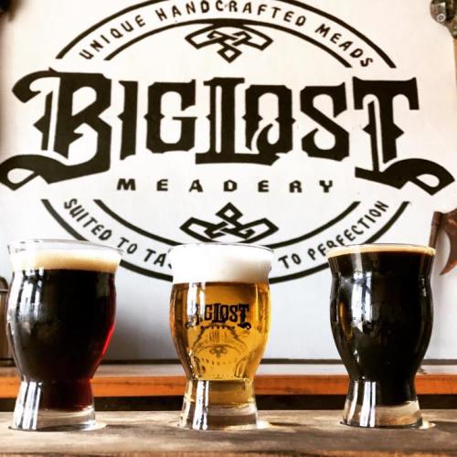 Three small glasses of mead in front of a "Big Lost Meadery" sign, showcasing unique handcrafted beverages.