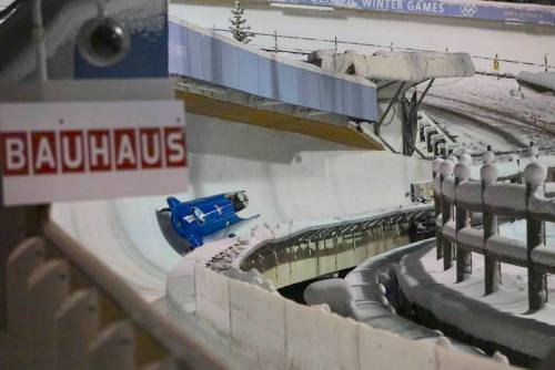 A blue bobsled racing down a snowy track at a winter sports venue, with a sign reading "BAUHAUS" in the foreground.