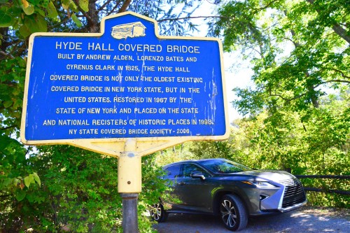 Sign for Hyde Hall Covered Bridge, built in 1825, with a car parked nearby and greenery in the background.