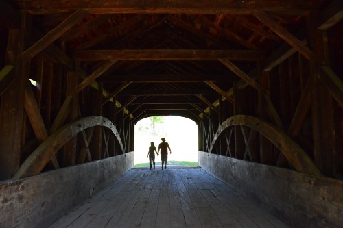 Silhouetted couple walking hand in hand through a wooden covered bridge, light streaming in from the exit.