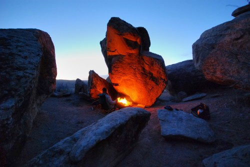 A campfire glowing between large rocks at dusk, with a clear blue sky in the background.