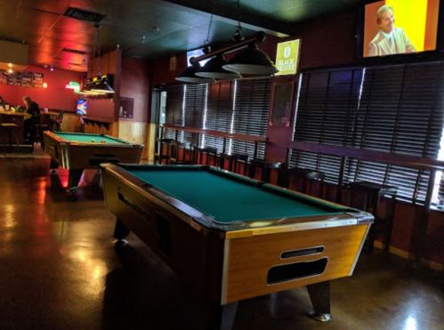 A dimly lit pool hall with two green-felt tables and a bar in the background, featuring a TV on the wall.