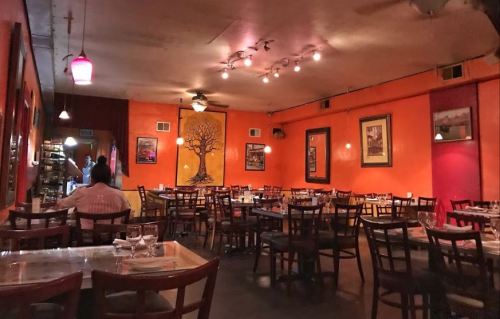 Cozy restaurant interior with orange walls, wooden tables, and a tree mural, featuring dim lighting and empty chairs.