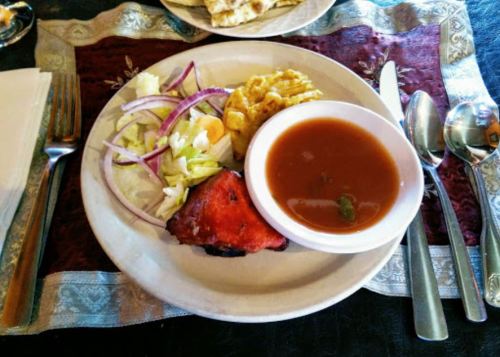 A plate of food featuring grilled chicken, salad, fried potatoes, and a bowl of soup, set on a decorative tablecloth.