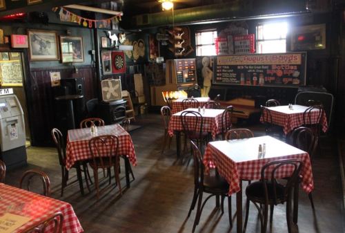 Cozy restaurant interior with checkered tablecloths, wooden chairs, and vintage decor, featuring a dartboard and a stage.