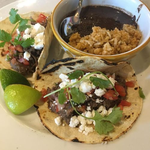 Two tacos filled with beef, topped with cheese and cilantro, served with rice and a bowl of black beans, garnished with lime.