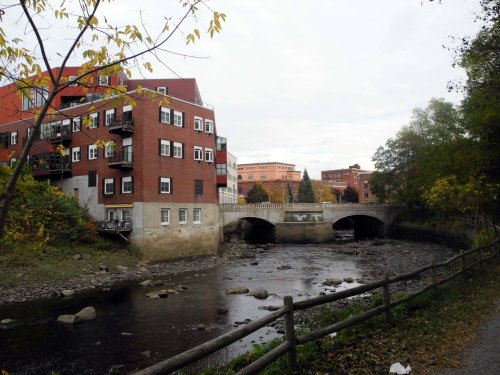 A scenic view of a river with a stone bridge, surrounded by autumn trees and red-brick buildings in the background.