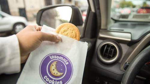 A hand holding a cookie from an Insomnia Cookies bag inside a car, with the steering wheel and dashboard visible.
