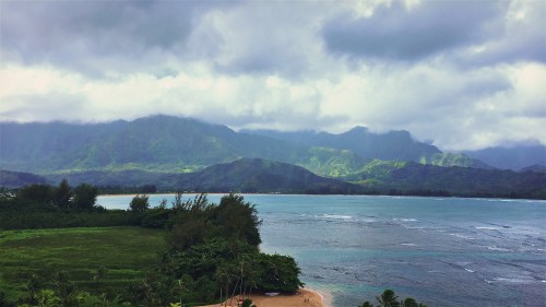 A scenic view of mountains and a calm ocean under a cloudy sky, with lush greenery along the shoreline.