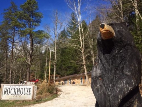 A large wooden bear sculpture stands near the Red River Rockhouse Cafe, surrounded by trees and a gravel path.