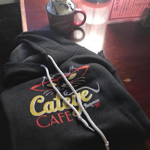 A black hoodie with a colorful logo of Cateye Cafe, next to a mug topped with whipped cream.