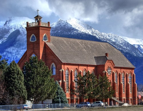 A red brick church with a cross on top, set against a backdrop of snow-capped mountains and a cloudy sky.