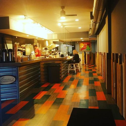 A cozy café interior with wooden accents, colorful flooring, and a customer sitting at a table while staff prepare food.