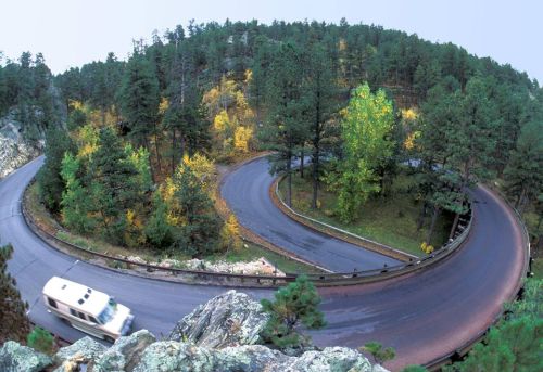 A winding road curves through a forested area with autumn foliage, as a vehicle drives along the scenic route.