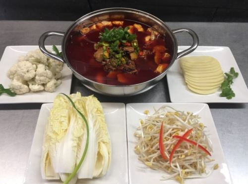 A spicy hot pot with tofu and meat, surrounded by fresh vegetables like cauliflower, lettuce, and bean sprouts.