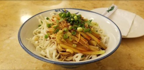 A bowl of noodles topped with green onions and vegetables, served on a textured surface with a napkin beside it.