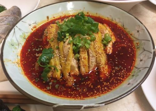 A bowl of spicy chicken dish topped with green onions and sesame seeds in a vibrant red sauce.