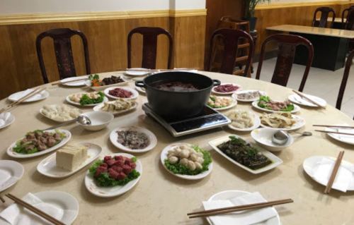 A round table set with various dishes and a hot pot in the center, surrounded by plates of meat, vegetables, and noodles.