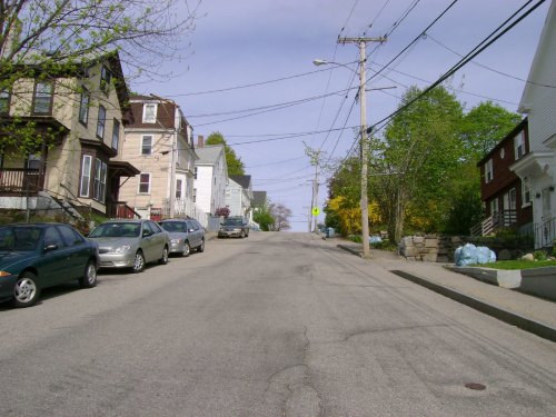A residential street lined with houses, parked cars, and trees, leading up a gentle hill under a cloudy sky.