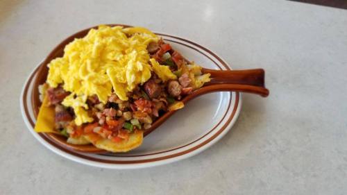 A brown dish filled with nachos topped with scrambled eggs, diced tomatoes, and sausage.
