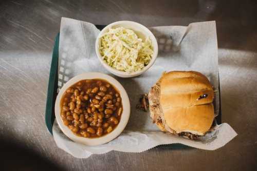 A tray with a pulled pork sandwich, a bowl of baked beans, and a small serving of coleslaw on a paper liner.