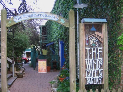 Entrance to The Winds Café & Bakery, featuring a wooden sign and lush greenery surrounding the pathway.